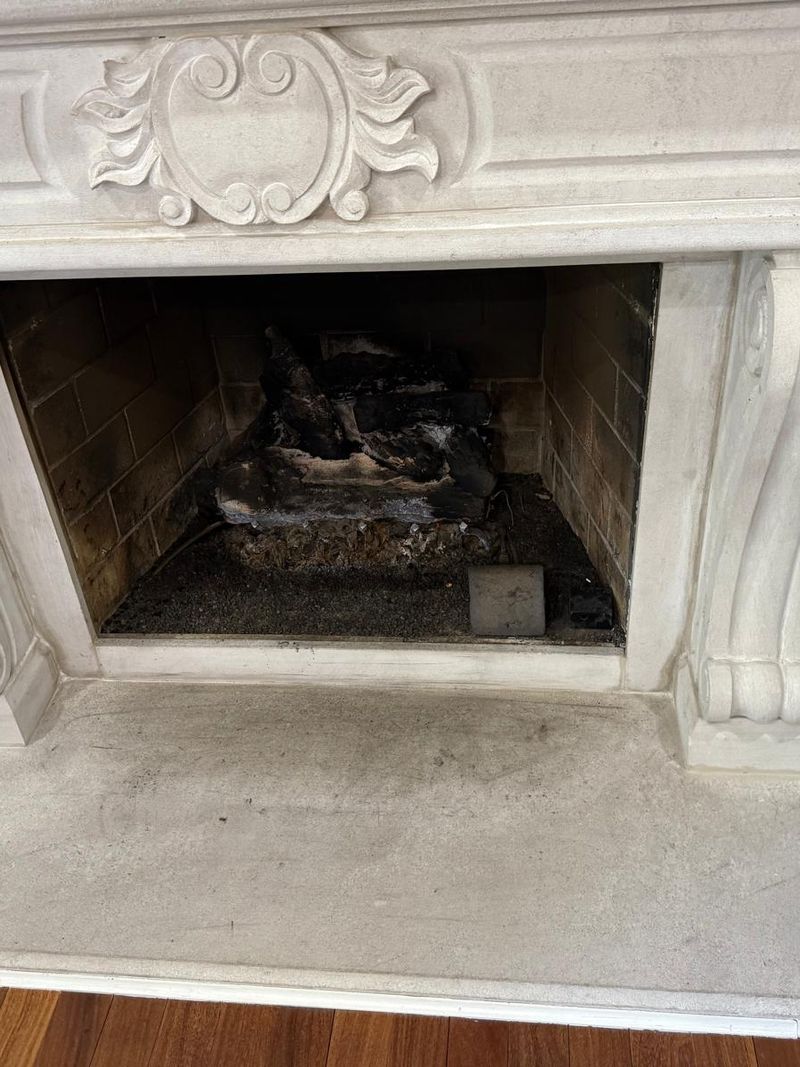 Carved stone fireplace mantel with scroll corbels and a sooty firebox holding charred gas logs