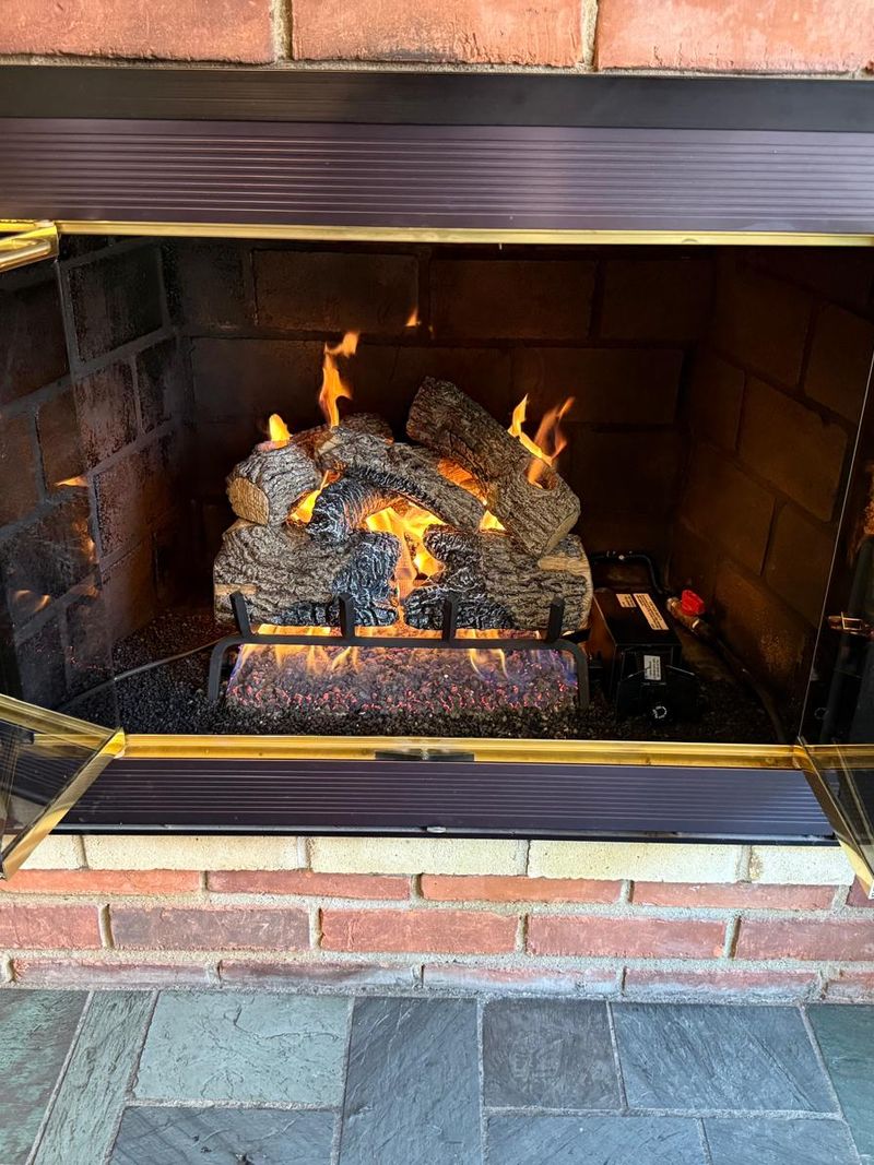 Gas log set burning with tall flames inside a red brick fireplace with open bifold glass doors and a slate hearth