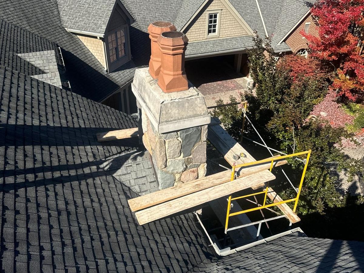Fieldstone chimney with a stone cap and two terra cotta chimney pots on a large gray-shingled house in the fall