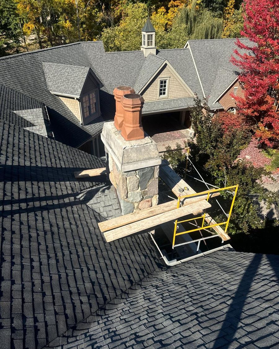 Fieldstone chimney with two terra cotta chimney pots beside a large house and autumn trees
