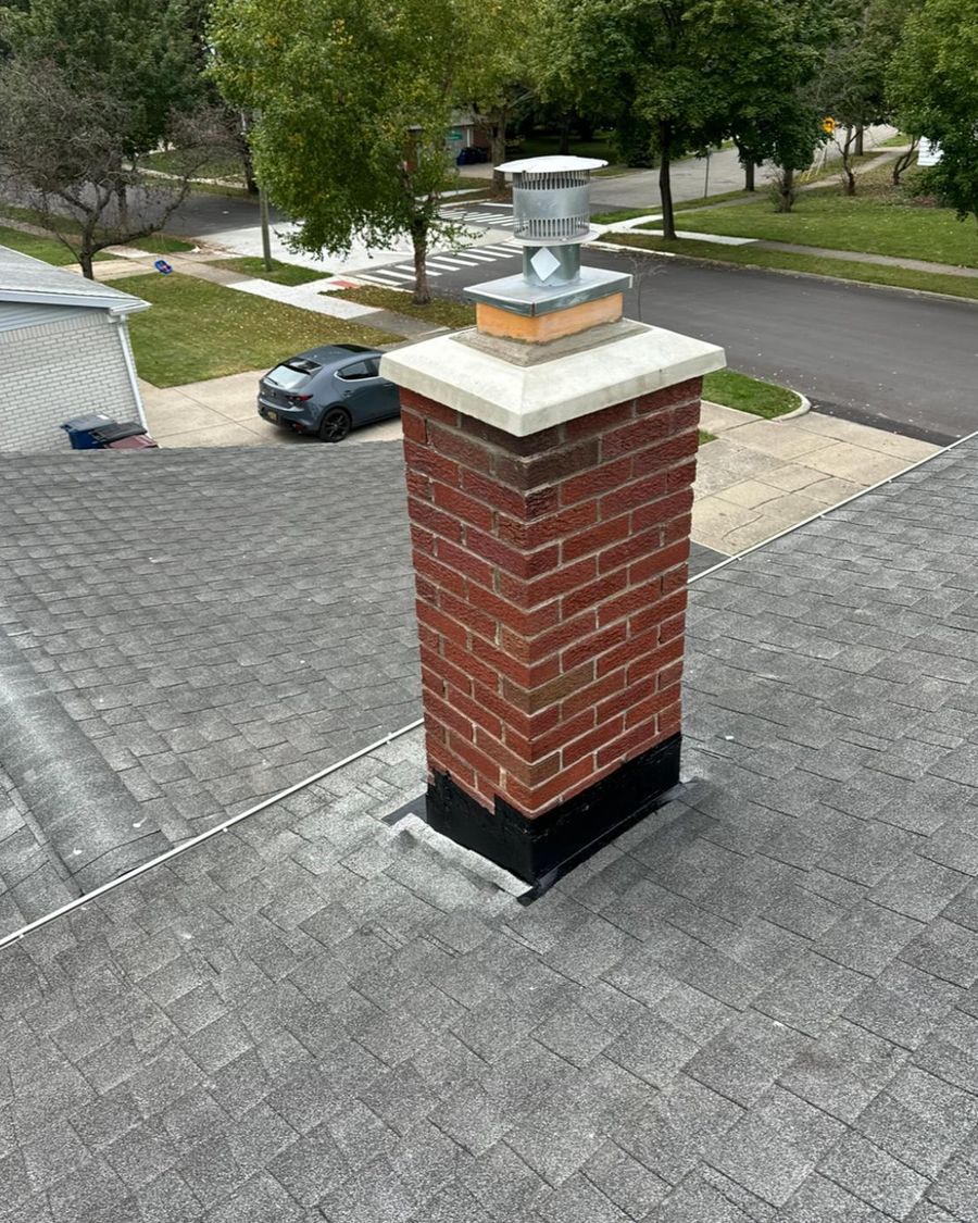 Red brick chimney with a white concrete crown and a stainless steel cap on a gray shingle roof