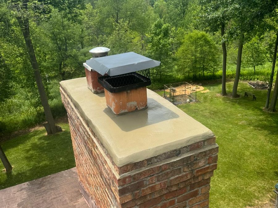 Red brick chimney with a smooth mortar crown and a black mesh cap above a wooded backyard