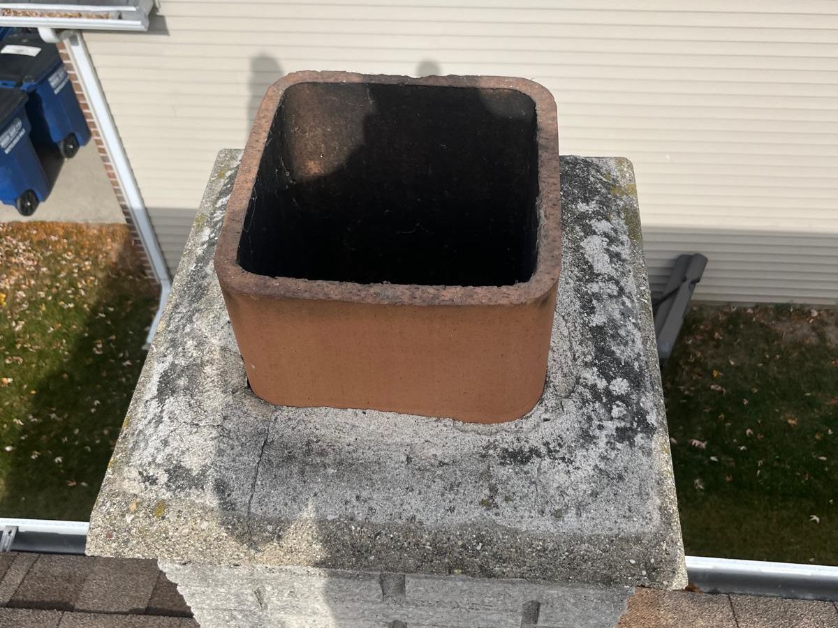 Concrete block chimney with one clay flue tile, a lichen-stained crown and no cap, above a brown shingled roof next to a sided house