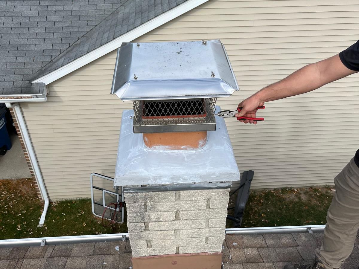 A gloved hand using pliers to fasten a stainless steel mesh chimney cap onto a clay flue above a white-coated crown