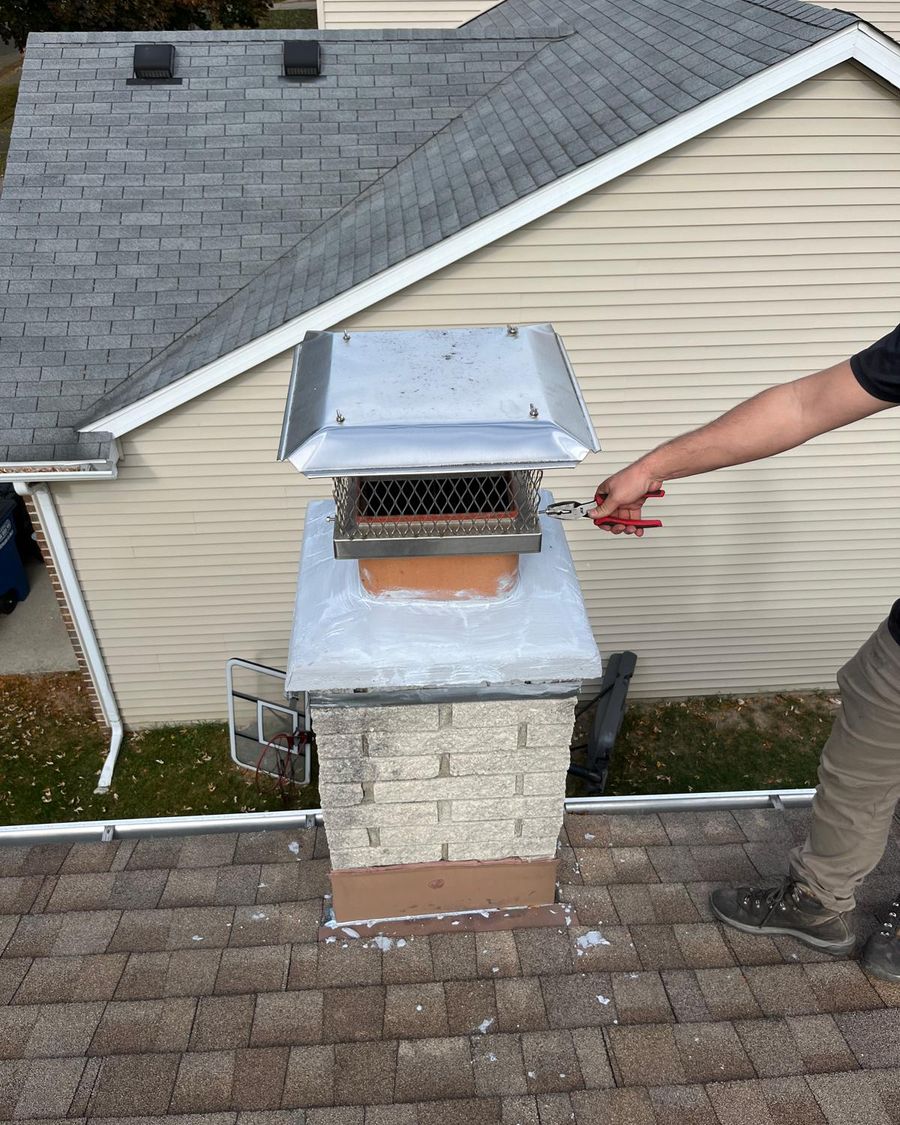 Hands using pliers to fasten a stainless steel chimney cap onto a flue above a white-coated crown