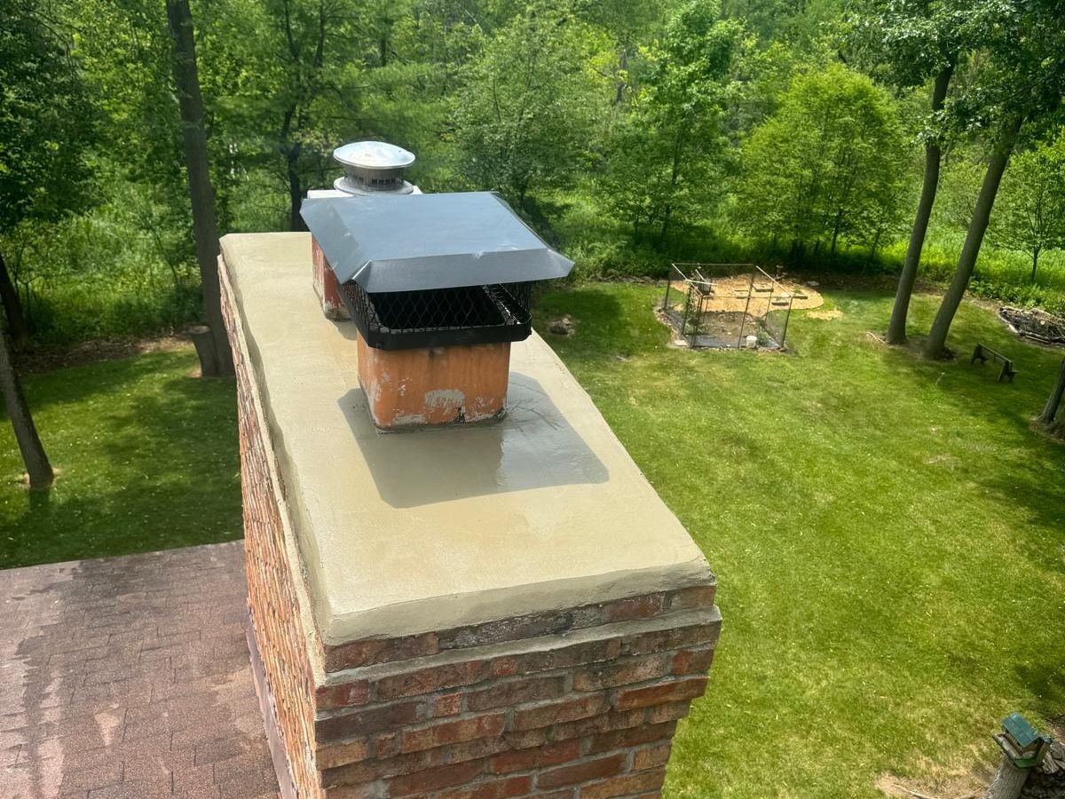 Tall red brick chimney with a smooth mortar crown and a black mesh cap above a tree-lined yard