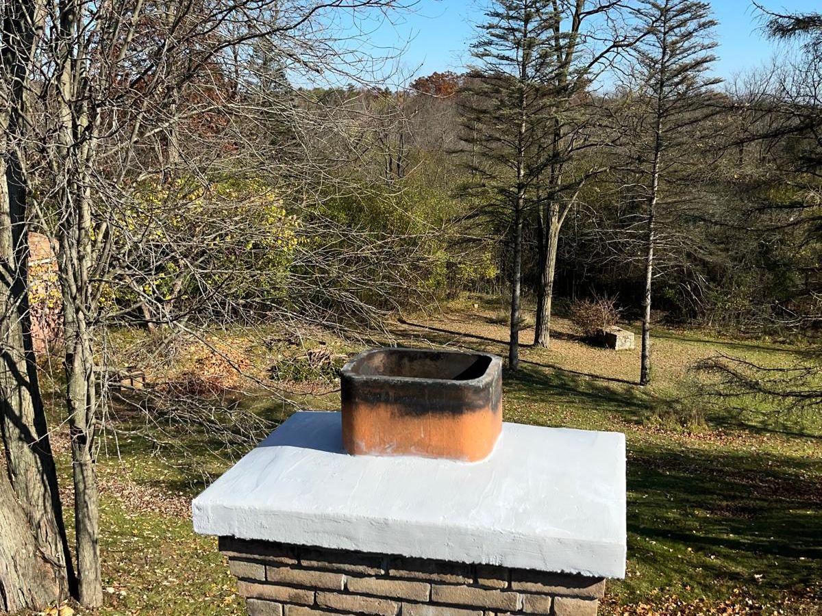 Tan brick chimney with a newly coated crown around a clay flue tile, with wooded land behind