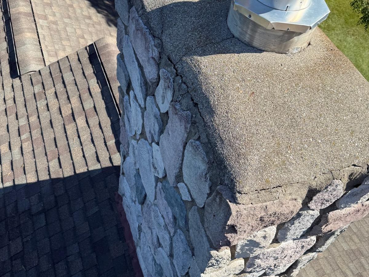 Fieldstone chimney with a weathered concrete crown cracked through near the edge and around a tall stainless steel vent pipe