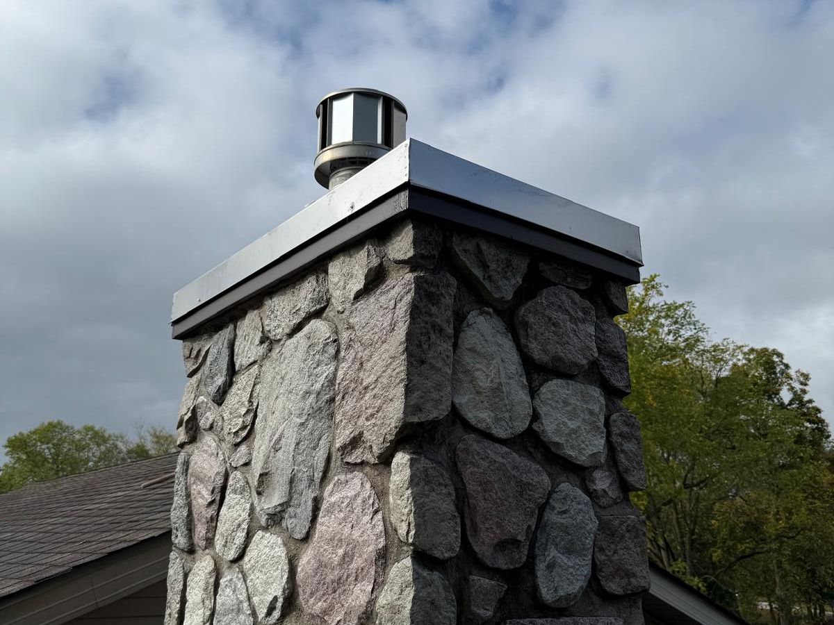 Fieldstone chimney with a stainless steel cover rising against a cloudy blue sky