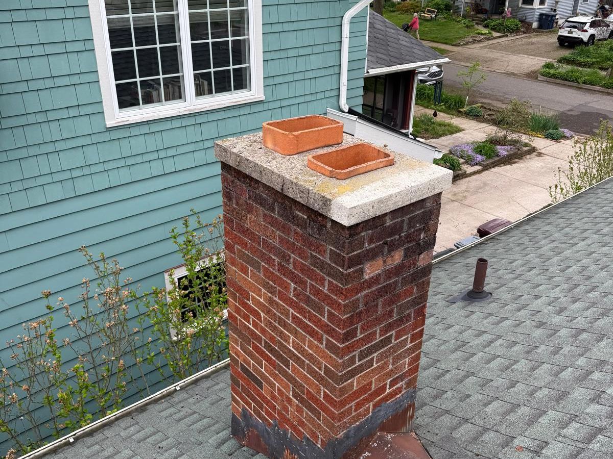 Red brick chimney with a lichen-covered crown, loose clay flue tiles and rusted base flashing beside a teal house