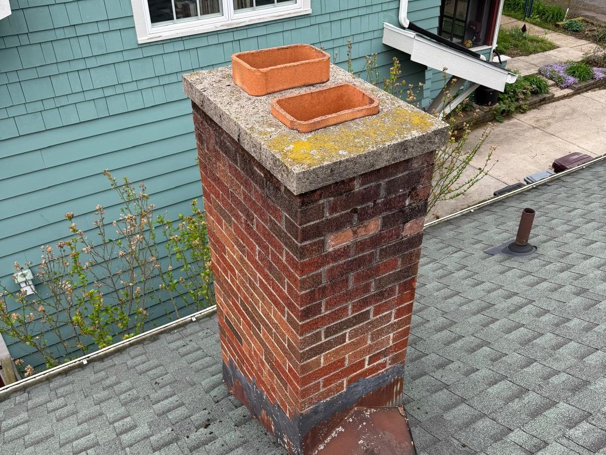 Red brick chimney with two clay flue tiles, lichen on the concrete crown and rusted flashing at the roof, beside a teal shingled wall
