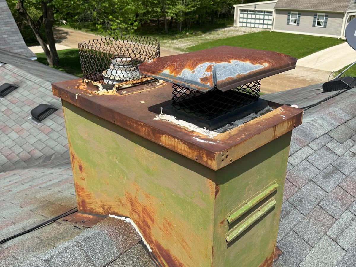 Metal chimney chase with peeling green paint, a rusted chase cover and a rusted cap with a mesh screen, on an asphalt shingle roof