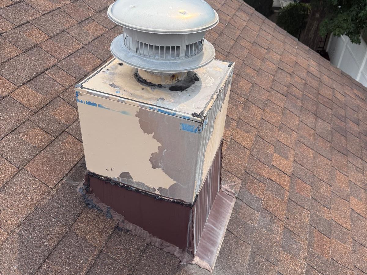Short metal chimney chase on a shingled roof with peeling paint, tar-sealed seams and a round metal vent cap