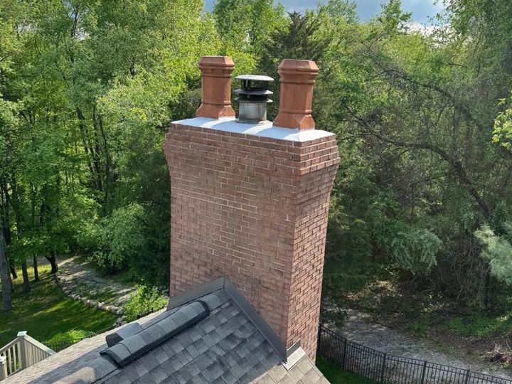 Red brick chimney with two terra cotta chimney pots, a stainless steel flue cap and a white crown, above a wooded backyard with an iron fence