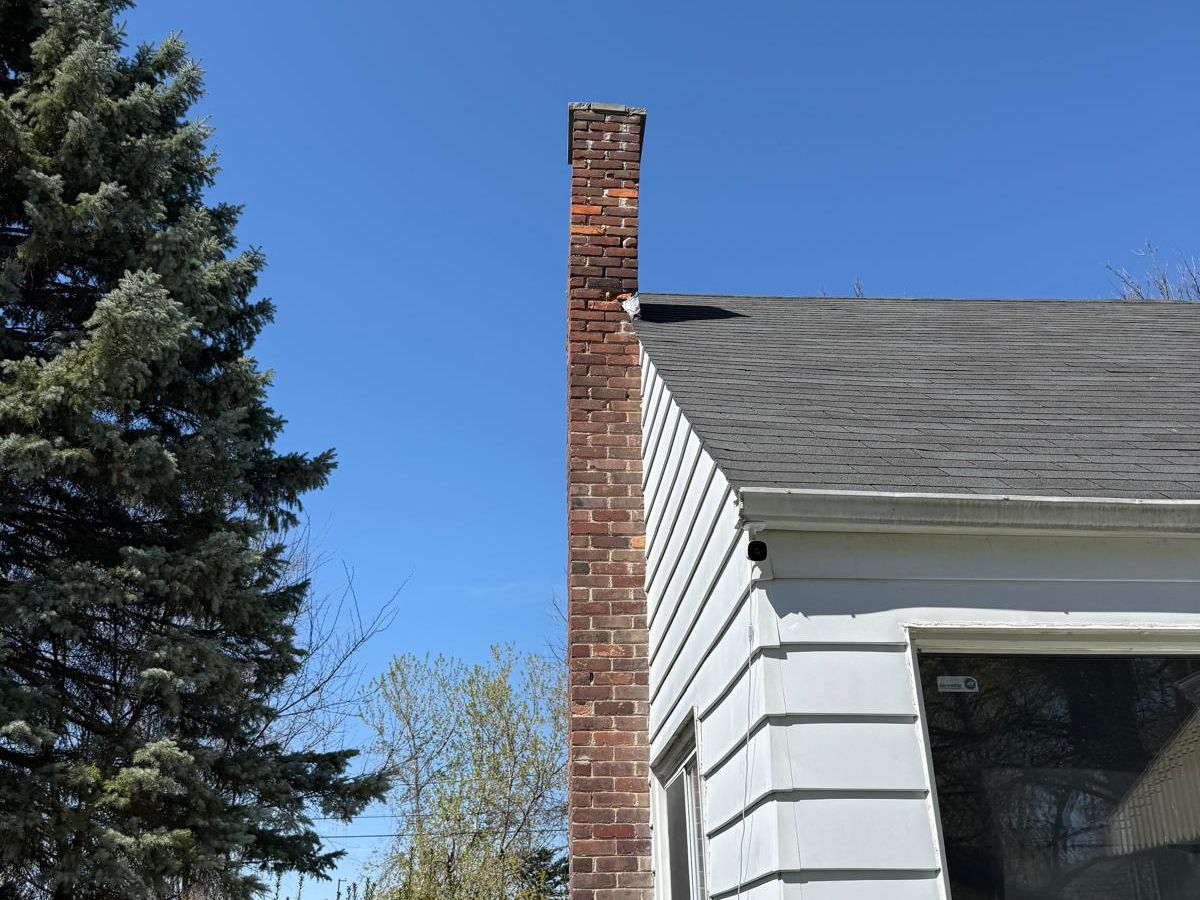 Tall, narrow brick chimney running up the side of a white-sided house under a clear blue sky
