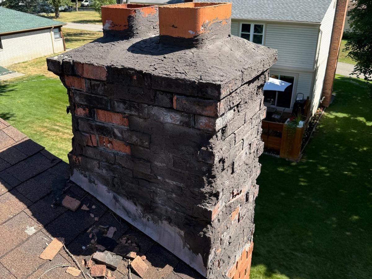 Brick chimney with roofing tar smeared over the crown and upper brick, crumbling brick faces, two clay flue tiles, and broken brick pieces on the roof